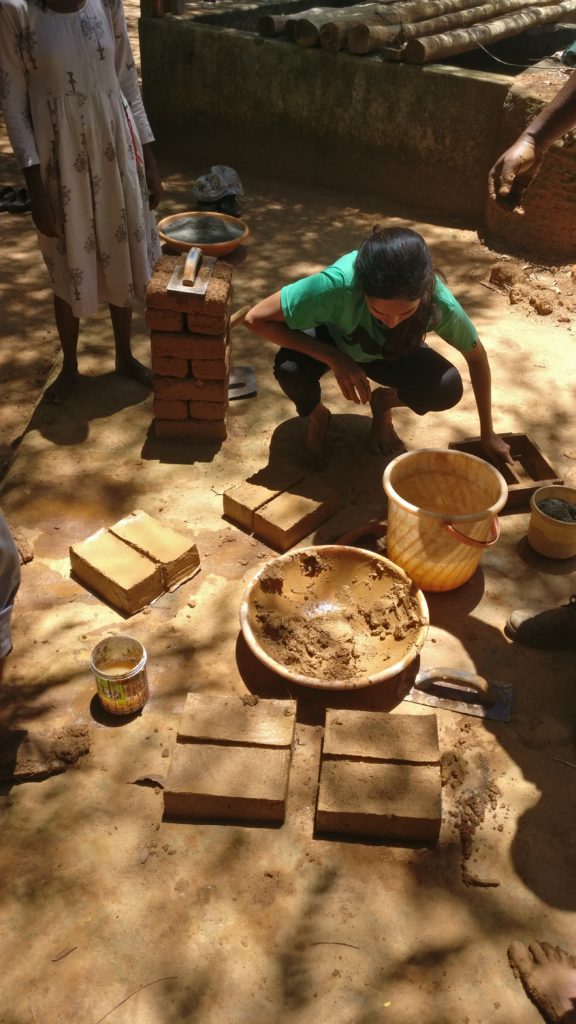 A girl is preparing mud bricks using a mould. A stack of bricks is shown. Another girl is standing by.