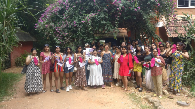 A group of girls standing in front of a mud house with trees all around.