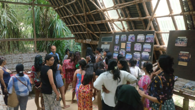 A few girls and a man standing under a bamboo hut with photographs on a notice board